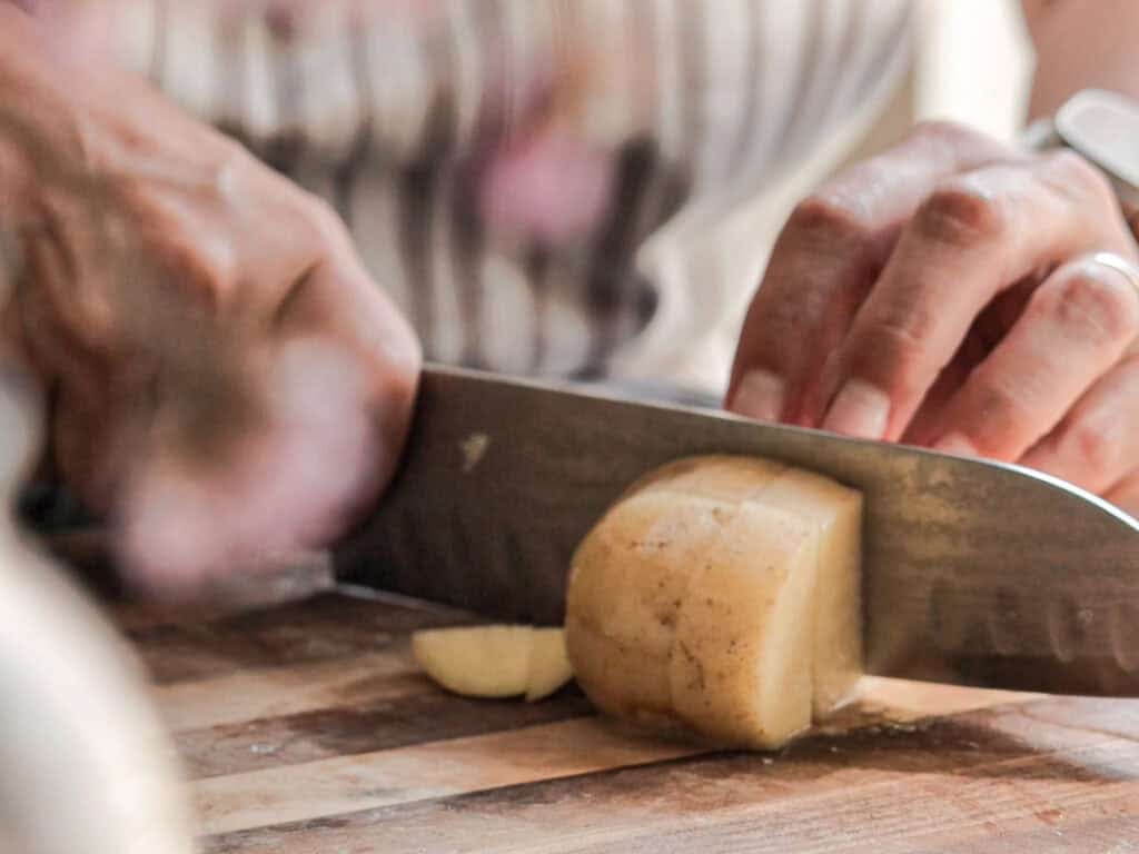 Close-up of hands slicing a potato with a large knife on a wooden cutting board. The person is wearing a striped apron, and the focus is on the knife and potato. The background is blurred.